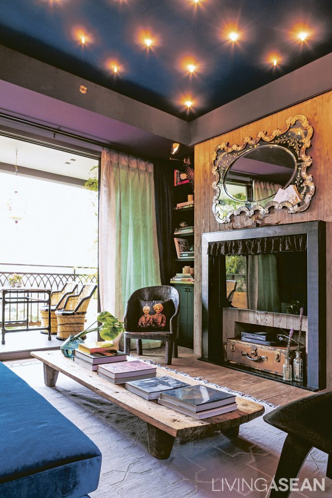 Dark colors alternate with paler ones, giving dimension to the "living room." The light gray wall is adorned with framed pictures. The teakwood floor is polished to a lighter shade, contrasting with the dark ceiling, and opens out onto the balcony, with its set of wicker chairs.