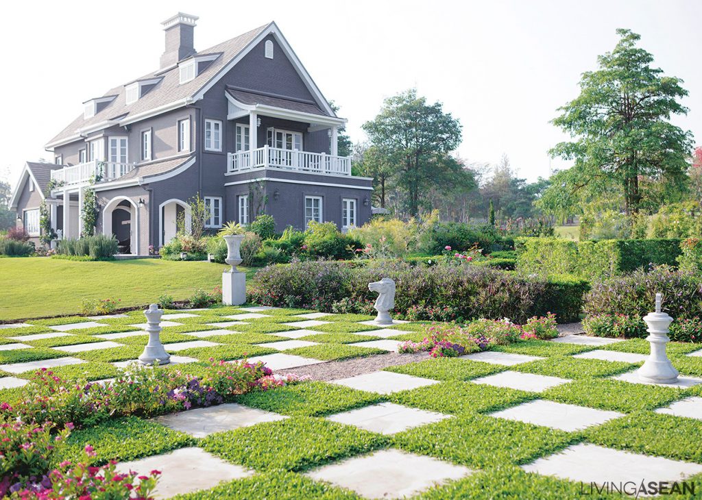 This grey house in the “English Country” style stands out in the middle of a European-style garden. Open lawns, variegated flowerbeds and flowering plants are spaced at intervals. 