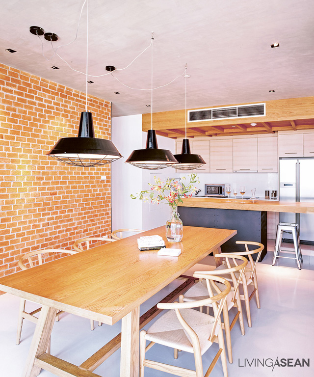 The elegant dining area with a long table connects the counter to the kitchen at the rear. Metal black lamps hang from the ceiling, contrasting with the warm colors of brick and wood