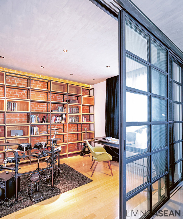 A floor-to-ceiling sliding door with safety glass panels in a black metal frame separates off the workspace and drum practice room. Artistry design is evident in the shelving all along the back wall, where the fancy brick shows through open spaces in the frame. 