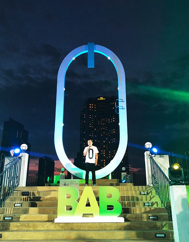 Sporting a “Zero” T-shirt with Elmgreen and Dragset autographs on it, Prof. Dr. Apinan Poshyananda, CEO and artistic director of the Bangkok Art Biennale 2018, gives an opening speech on the riverside platform aglow under spotlights.